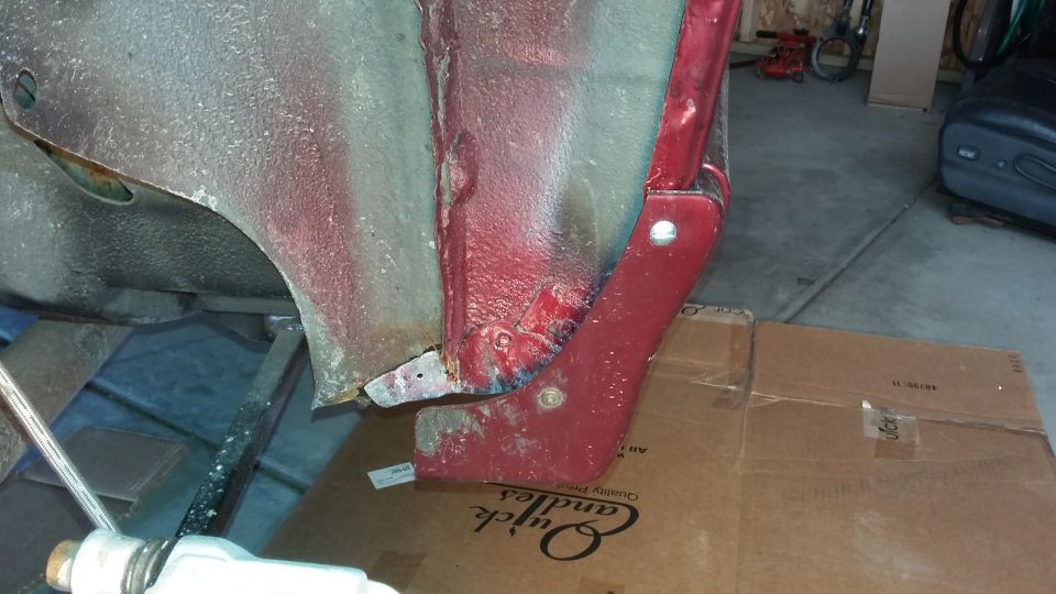 Lower corner of a red body panel with rust and a metal repair patch in a garage
