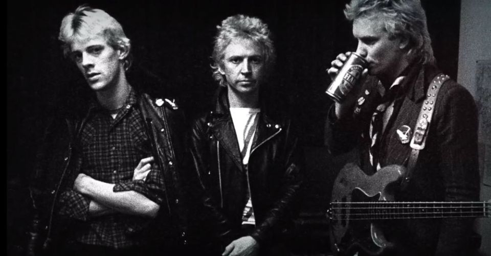 Three men in leather jackets, one drinking from a can and holding a guitar