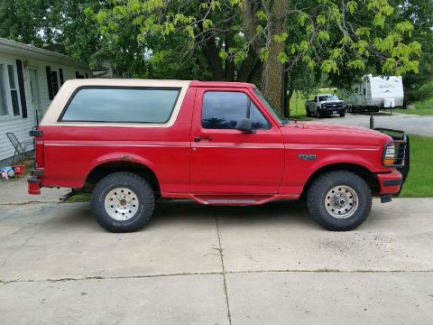 1996 Ford Bronco - A red Ford Bronco with a tan top is parked on a concrete driveway, with another vehicle and a trailer in the background.