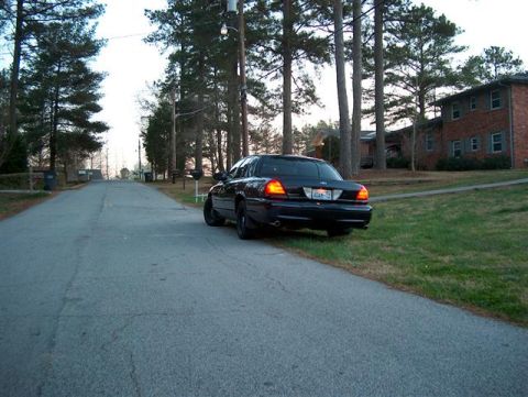 2001 Ford Crown Victoria - A dark-colored Ford Crown Victoria is parked on the grass next to a paved road in a suburban neighborhood.