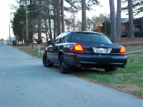 2001 Ford Crown Victoria - The rear of a dark blue 2001 Ford Crown Victoria is visible from a low angle, parked on the shoulder of a paved road.