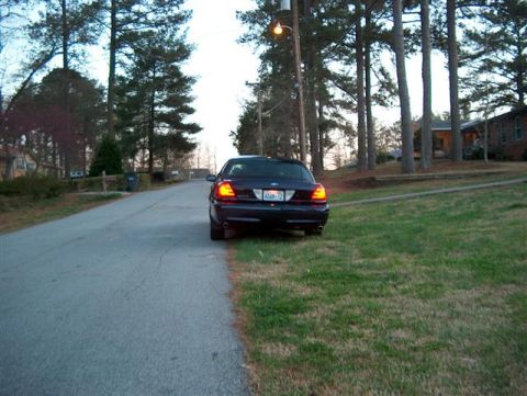 2001 Ford Crown Victoria - A dark sedan is parked on the side of a road in a residential area with trees and houses in the background.
