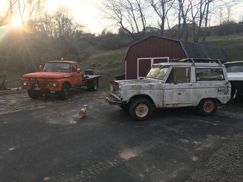 1967 Ford Bronco - Two vehicles, a white Ford Bronco and an orange flatbed truck, are parked in a driveway next to a red barn.