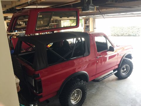 1994 Ford Bronco - A red Ford Bronco is parked in a garage with its top removed, and another Bronco top is suspended overhead.