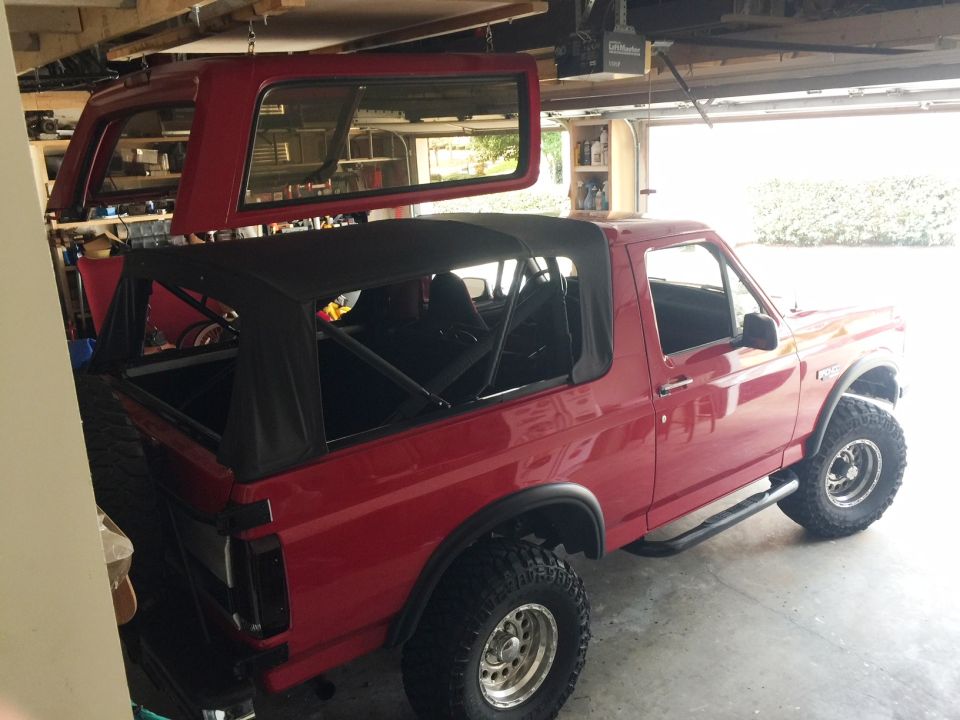 Red Ford Bronco with soft top removed, another red Bronco top section hanging above it in a garage