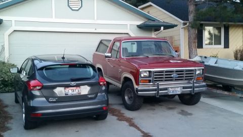 1985 Ford Bronco - A red 1985 Ford Bronco is parked in a driveway next to a modern hatchback and a boat on a trailer.