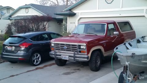 1985 Ford Bronco - A maroon 1980s Ford Bronco is parked in a driveway next to a dark gray Ford Focus. A small aluminum boat on a trailer is parked next to the…