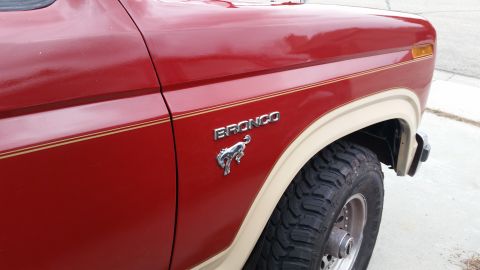 1985 Ford Bronco - Close-up view of the front fender and door of a red Ford Bronco. The vehicle has a tan trim line and a chrome Bronco emblem with a horse gr…