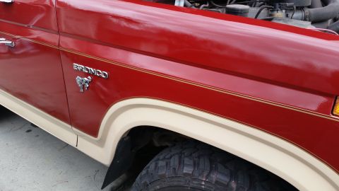 1985 Ford Bronco - Close-up side view of a red Ford Bronco fender with a tan lower trim and a chrome Bronco emblem. The vehicle has gold pinstriping along the…