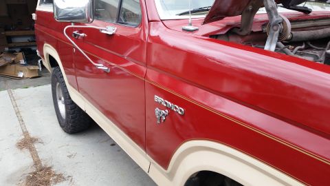 1985 Ford Bronco - Close-up view of the driver's side front fender and door of a red and tan Ford Bronco. The vehicle has chrome trim and a "Bronco" emblem wi…