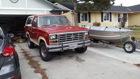 1985 Ford Bronco - A red 1980s Ford Bronco is parked in a driveway next to a boat on a trailer. A dark gray car is partially visible on the left side of the i…
