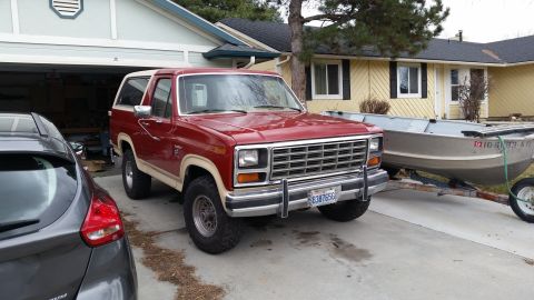 1985 Ford Bronco - A red and tan 1980s Ford Bronco is parked in a driveway next to a dark gray hatchback. A boat on a trailer is visible behind the Bronco.