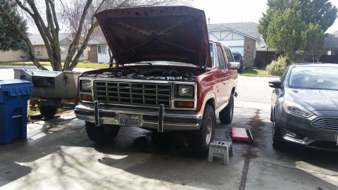 1985 Ford Bronco - The front end of a red and white 1980s Ford Bronco is shown with its hood open, parked in a driveway next to a dark gray Ford Focus. A blue…