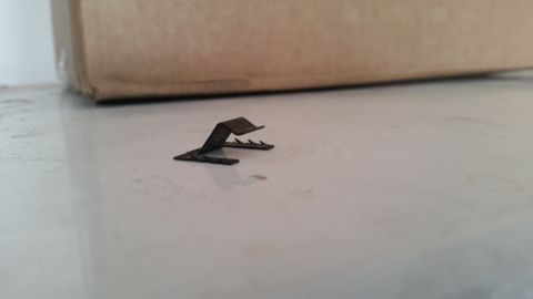 1985 Ford Bronco - A small, dark metal clip with sharp teeth rests on a reflective white surface. A portion of a brown cardboard box is visible in the backgro…
