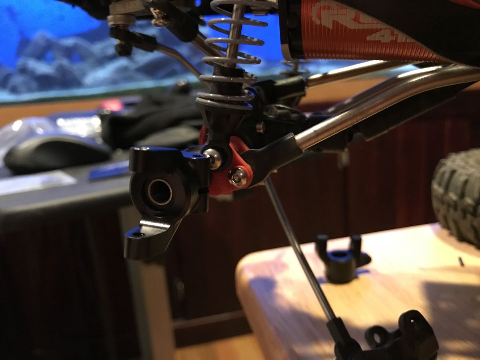Close-up of RC car suspension and steering components, black knuckle, silver arms, red accent.