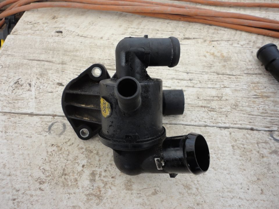 Black plastic thermostat housing with hose connections and a yellow sticker.