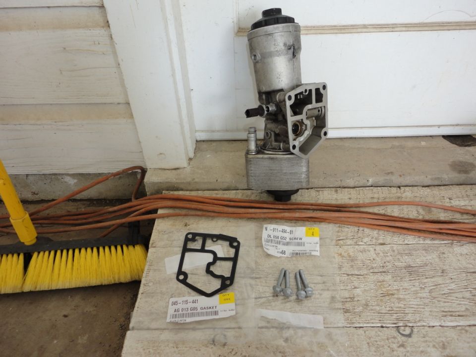 Disassembled oil filter housing, gasket, and screws on a wooden surface.
