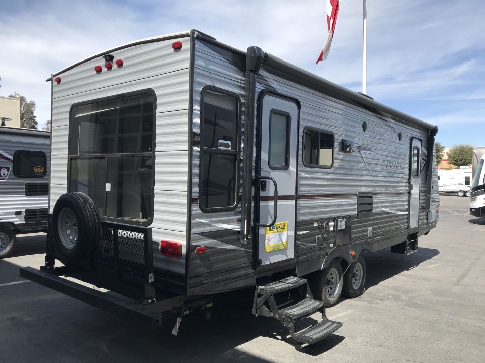 Grey and black travel trailer with spare tire on rear rack, parked outdoors.