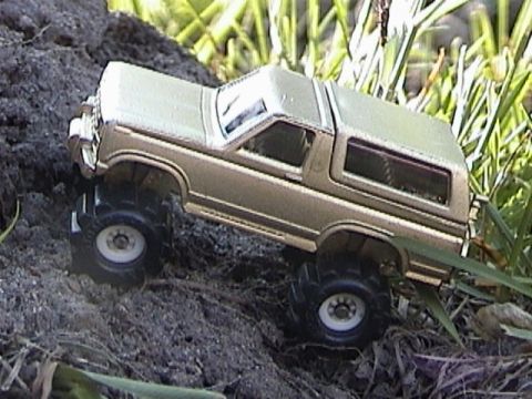 1979 Ford Bronco - A gold-and-white two-tone die-cast Ford Bronco posed outdoors on a mound of dark soil beside grass, shot from a high three-quarter rear ang…