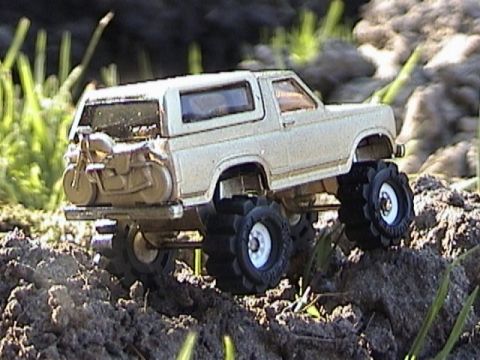 1979 Ford Bronco - A gold/tan die-cast full-size Bronco-style SUV toy photographed outdoors in three-quarter rear view, posed on a mound of dark dirt with gra…