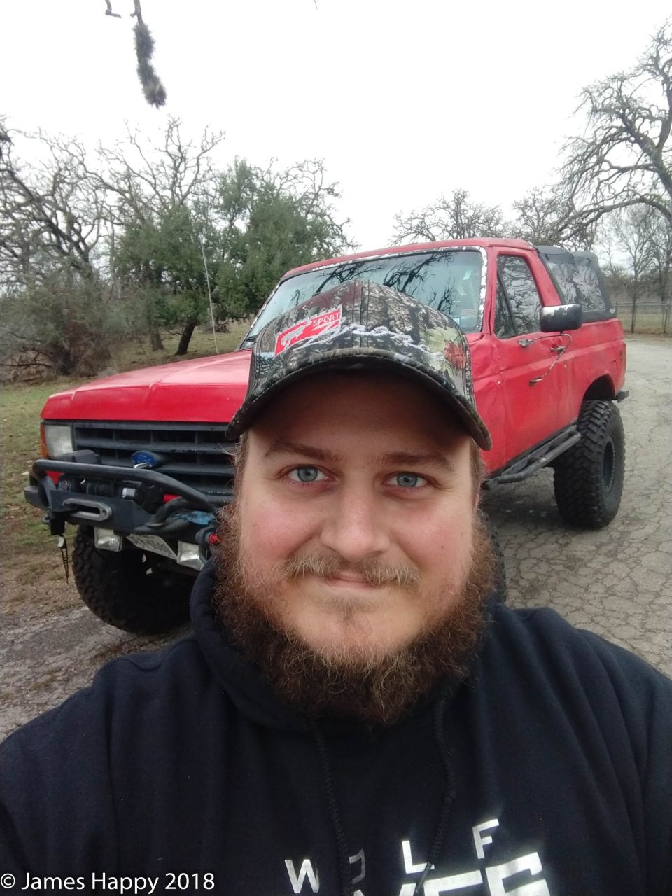 Man wearing a camo hat and black hoodie stands in front of a red SUV with off-road tires