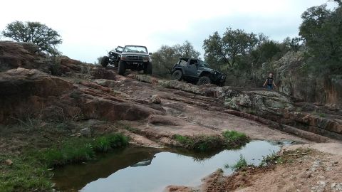 1995 Ford Bronco - Two off-road vehicles navigate a challenging rocky terrain with a small pool of water in the foreground.