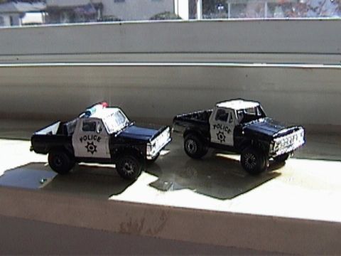 1979 Ford Bronco - Two black-and-white police-liveried die-cast Ford trucks posed side by side on a sunlit window sill, both wearing "POLICE" door lettering w…