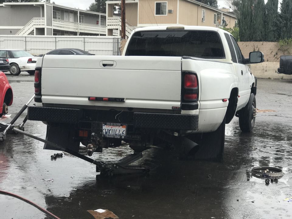 White pickup truck with dual rear wheels, lifted by jacks, with a diamond plate bumper and a license plate reading SHKULL46