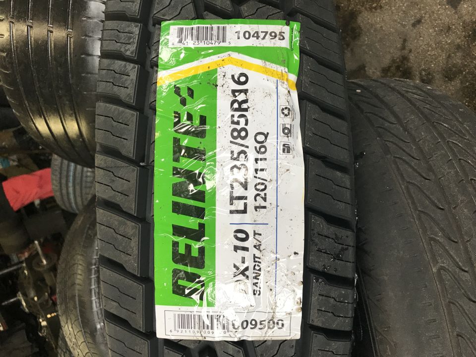 New tire with green and white label showing brand RELIANTEX, model X-10 LT235/85R16, load index 120/116Q, and type SANDIT A/T