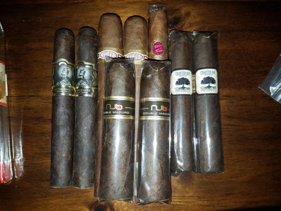 Several cigars on a wooden surface, some in plastic packaging with labels like "NUB DOUBLE MADURO" and "CHARTER OAK"