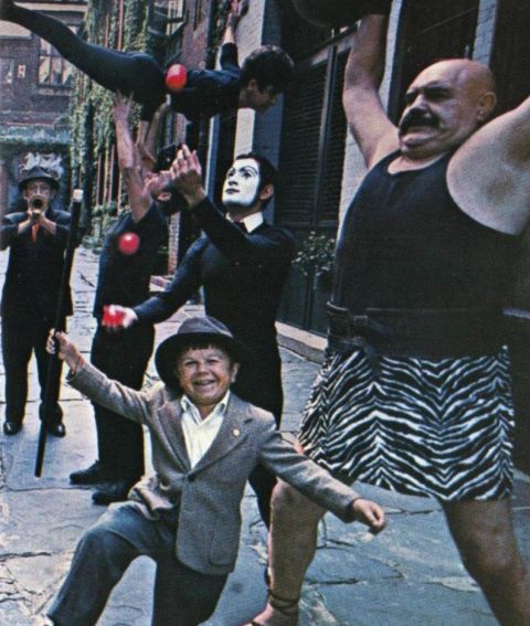 1967 Ford Custom Wagon - A group of circus performers and a child are gathered in a street, with a strongman lifting a weight overhead.