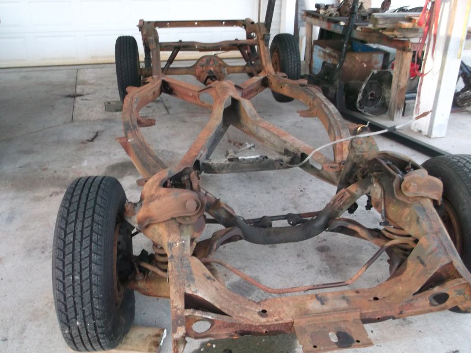 Rusty bare car chassis on four wheels sitting on a driveway in front of a garage during restoration