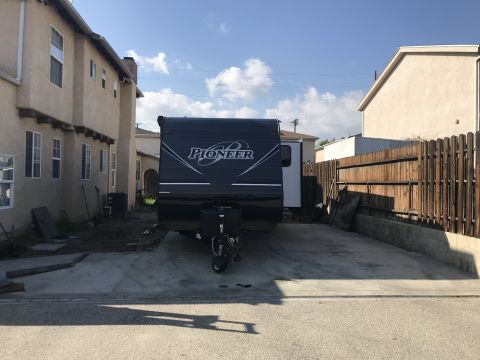 2018 Yugo GV - A Pioneer travel trailer is parked in a residential driveway between two houses.
