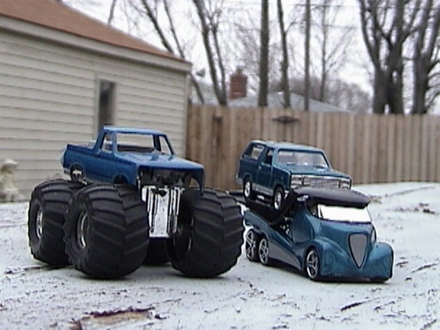 Toy monster truck, toy SUV on toy car carrier, and toy futuristic truck in snowy yard
