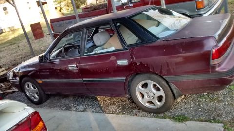 1994 Subaru Legacy - This maroon 1994 Subaru Legacy sedan shows significant wear, including peeling paint and damage to the front fender.