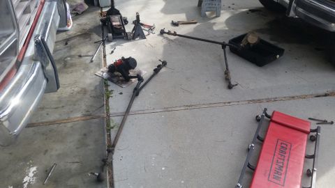 1985 Ford Bronco - Disassembled steering linkage and gearbox components are laid out on a concrete surface, with tools and a creeper nearby.