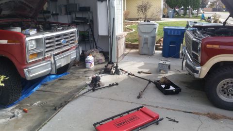 1985 Ford Bronco - Two Ford trucks are parked in a driveway, undergoing repairs with various parts laid out on the ground.