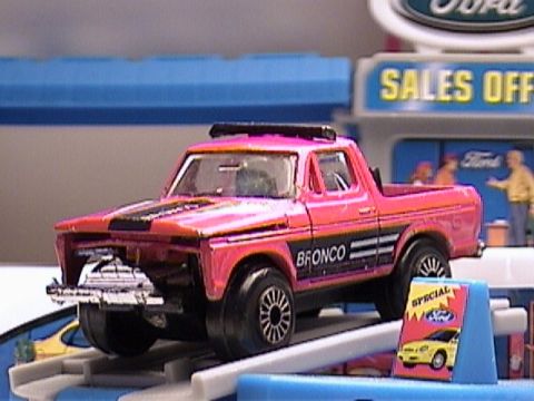 1979 Ford Bronco - A pink toy pickup truck is displayed on a blue ramp in front of a Ford dealership playset.