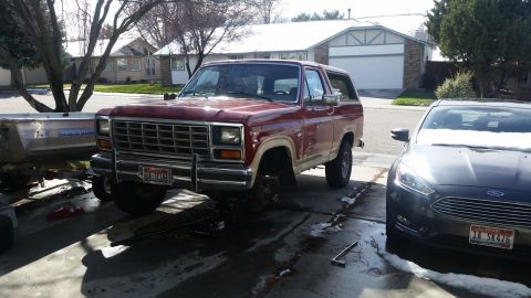 1985 Ford Bronco - A red Ford Bronco from the early 1980s is parked in a driveway, undergoing maintenance, with a boat trailer and another vehicle nearby.