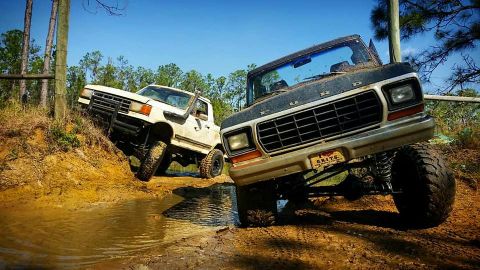 1988 Ford Bronco - Two off-road trucks are shown tackling a muddy terrain on a sunny day.