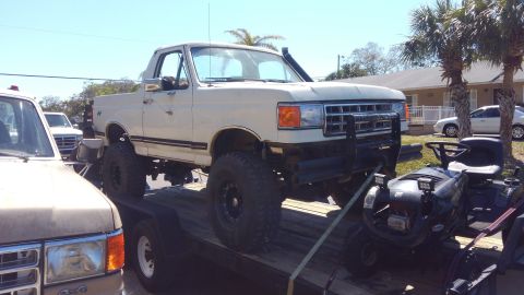 1988 Ford Bronco - A white pickup truck is loaded onto a trailer, accompanied by a riding mower and another vehicle.