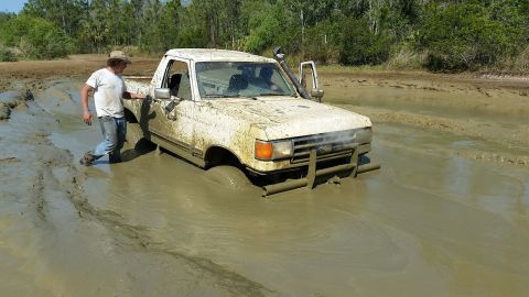 1988 Ford Bronco - Light-colored pickup truck buried to the doors in a muddy off-road pit while a man stands at the front fender.