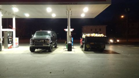 1996 Ford Bronco - Two vehicles, a dark van and a dump truck loaded with goods, are parked at a gas station under the canopy lights at night.
