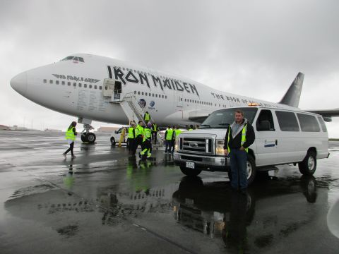 1996 Ford Bronco - A man stands next to a van on a wet airport tarmac with a Boeing 747 airplane, branded 'Iron Maiden', in the background.