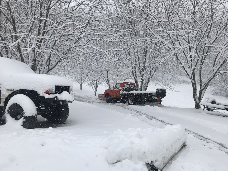 Orange pickup truck with a snow plow attachment in a snowy driveway. Another vehicle is partially visible on the left