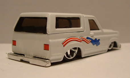 1979 Ford Bronco - White die-cast toy Ford Bronco slammed to the ground, photographed from the rear three-quarter against a plain gray backdrop. The custom mo…