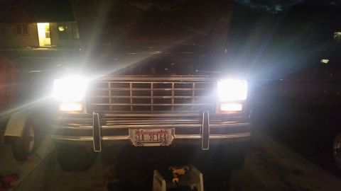 1985 Ford Bronco - A dark Ford Bronco is parked at night, its bright headlights illuminating the front grille and bumper.