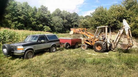 1994 Ford Bronco - A two-tone Ford Bronco tows a trailer carrying a backhoe in a rural setting with trees and a cornfield.