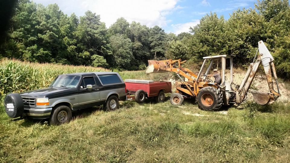 1994 Ford Bronco parked next to a trailer with a backhoe loader in a grassy field