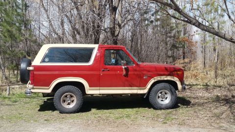 1985 Ford Bronco - This two-tone Ford Bronco is parked outdoors amidst a wooded area.